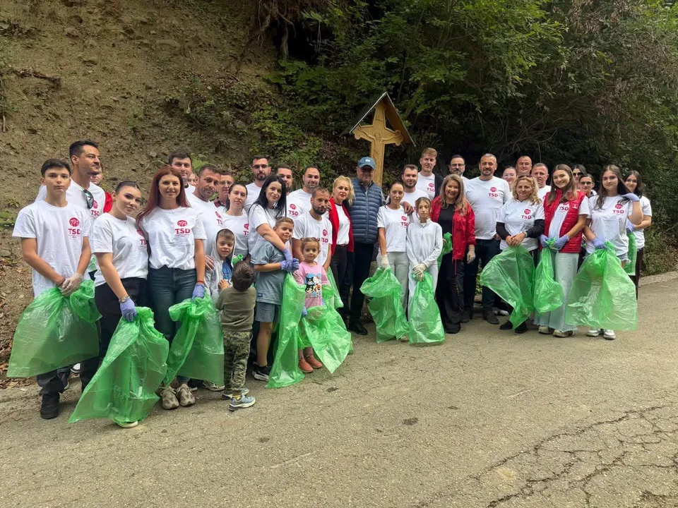 Un grup de oameni stă pe un drum asfaltat, purtând tricouri albe și ținând saci verzi de plastic.