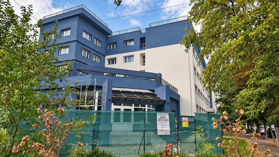 O clădire modernă cu fațadă albastră și albă este parțial acoperită de copaci și un gard de tip plasă verde în prim-plan.