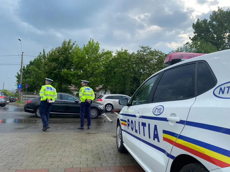 Doi polițiști în uniforme galben-verzui stau pe o stradă lângă un autoturism negru și o mașină de poliție albă.