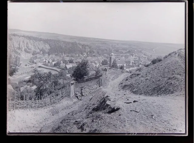 O fotografie alb-negru prezintă o vedere panoramică asupra unui sat situat într-o vale, cu dealuri împădurite și un teren accidentat în prim-plan.