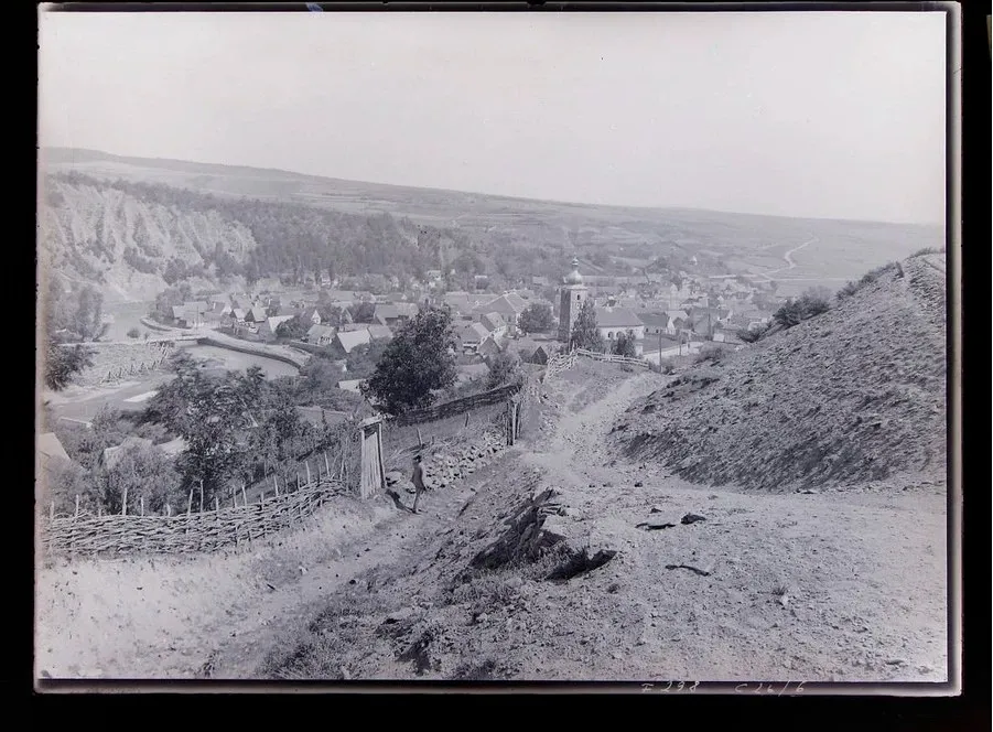 O fotografie alb-negru prezintă o vedere panoramică asupra unui sat situat într-o vale, cu dealuri împădurite și un teren accidentat în prim-plan.