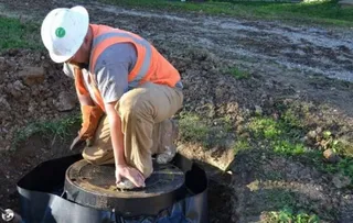 Un bărbat cu cască albă și vestă reflectorizantă orange este ghemuit lângă un capac de manhole într-o groapă de pământ.