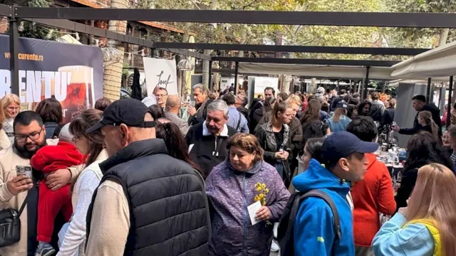 O mulțime de oameni stă în picioare într-un spațiu deschis cu umbră, lângă mese și standuri acoperite de prețuri.