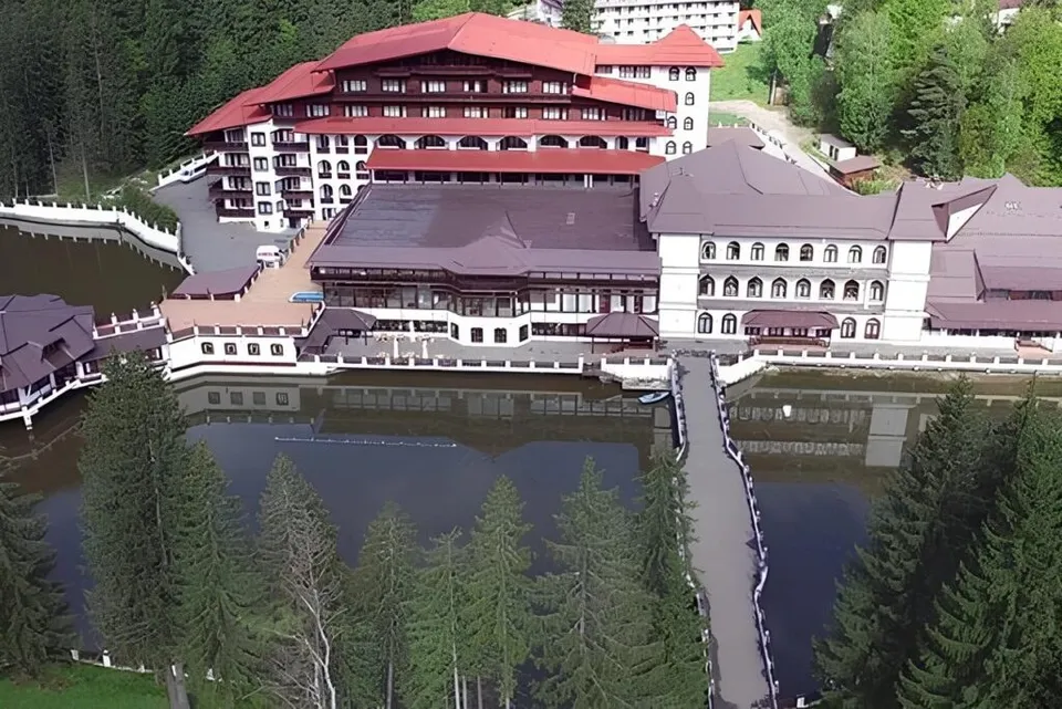 O clădire mare cu acoperiș roșu și maro este situată lângă un lac, înconjurată de copaci verzi.