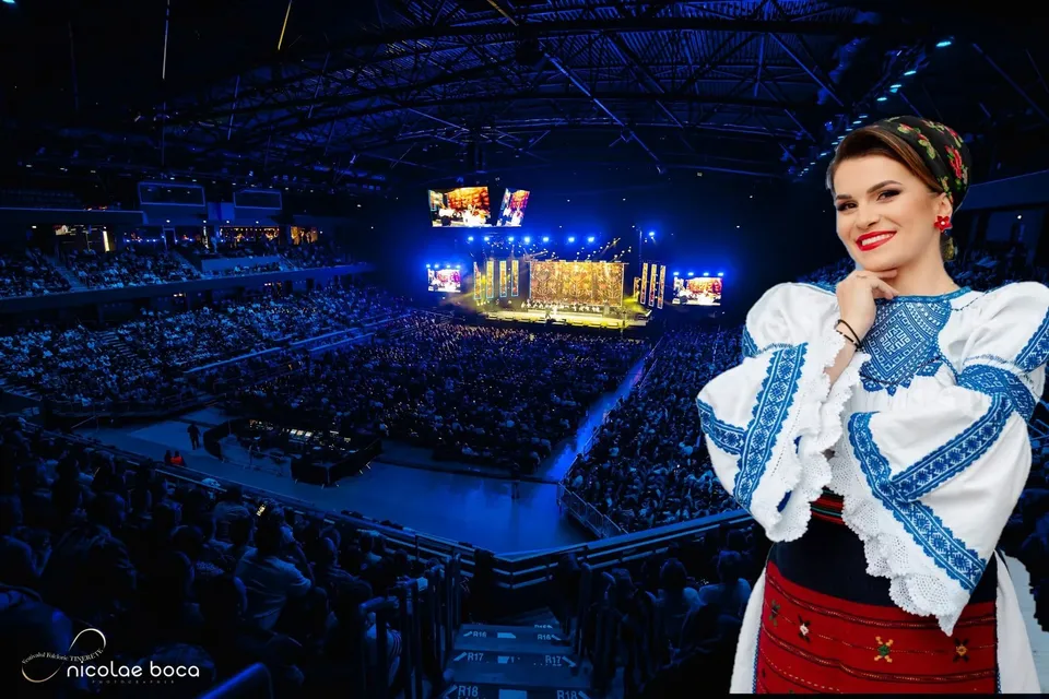 O femeie zâmbind poartă o cămașă tradițională albă cu broderii albastre, fiind plasată în fața unei săli de sport cu un public numeros și lumină albastră.