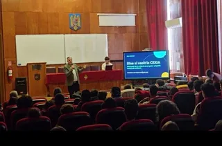 Un bărbat stă în picioare în fața unui ecran și prezintă ceva unei audiențe așezate în scaune într-o sală cu pereți din lemn.