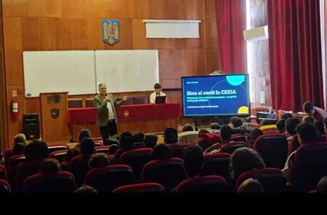 Un bărbat stă în picioare în fața unui ecran și prezintă ceva unei audiențe așezate în scaune într-o sală cu pereți din lemn.