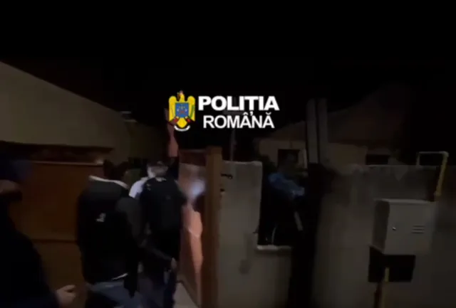 Patru persoane în uniformă întunecată stau lângă un gard și o poartă de lemn într-un decor nocturn.