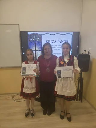 Două fete și o femeie stau în picioare în fața unui ecran care afișează un text, purtând certificate în mâini.