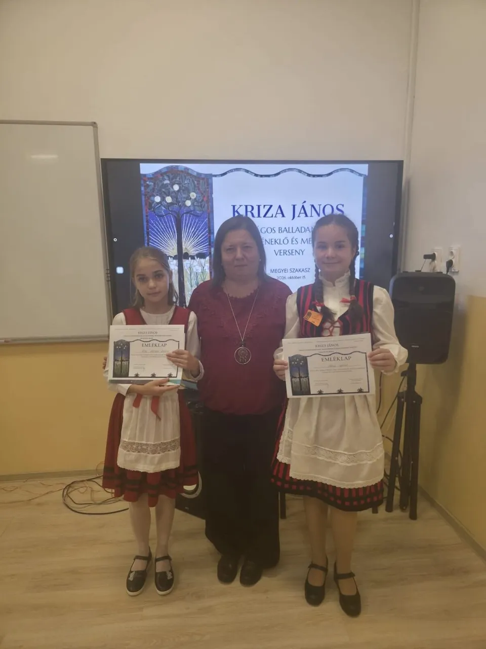 Două fete și o femeie stau în picioare în fața unui ecran care afișează un text, purtând certificate în mâini.