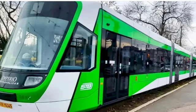 Un tramvai verde și alb cu inscripția STB circulă pe șine lângă un trotuar și copaci fără frunze.
