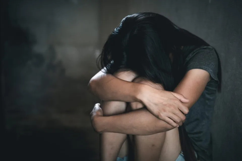 O persoană cu părul lung stă ghemuită cu capul plecat și brațele încrucișate în jurul genunchilor într-un decor întunecat.
