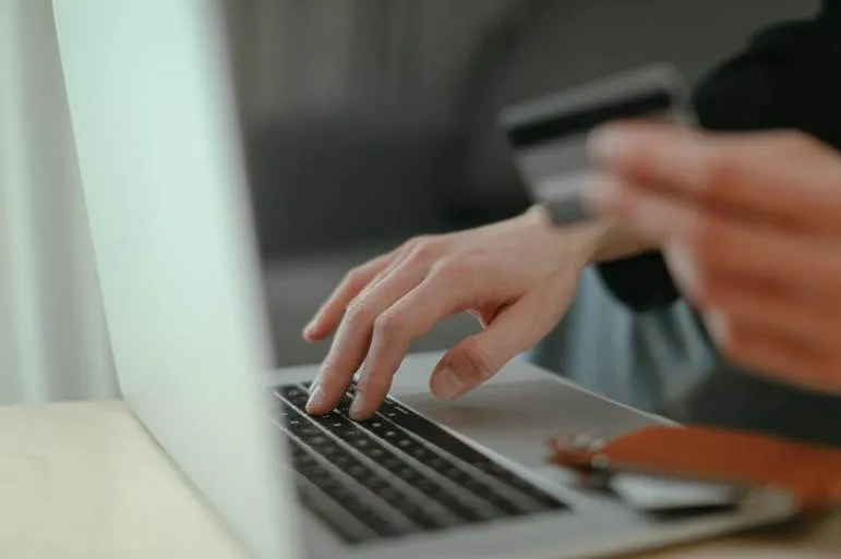 O persoană ține un card bancar în apropierea unui laptop deschis aflat pe o masă de lemn.