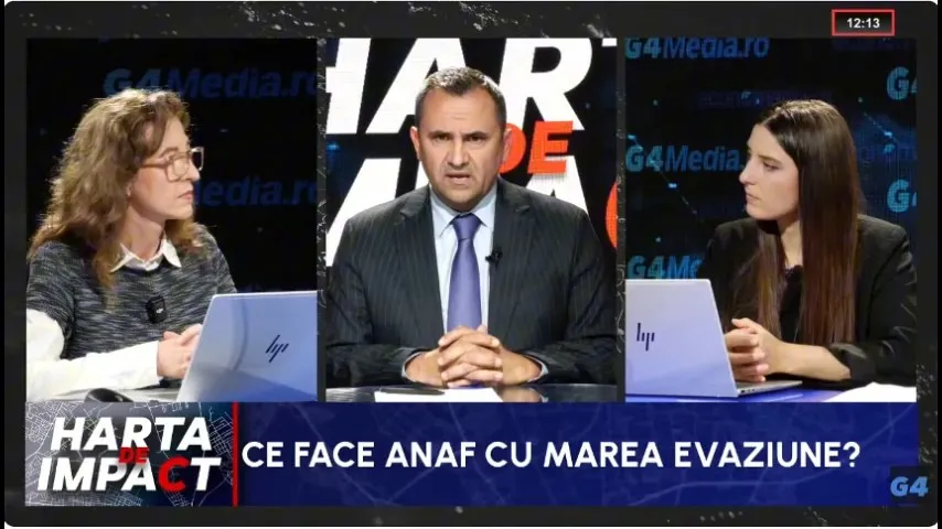 Trei persoane stau la o masă cu laptopuri deschise, în timp ce un bărbat în costum și cravată vorbește în fața unui fundal grafic.