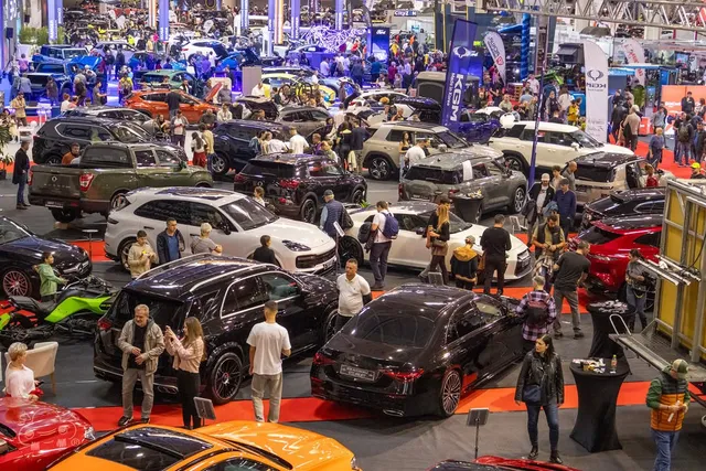 Mulți oameni vizitează un expoziție de mașini într-o hală mare cu diverse modele de autoturisme colorate și standuri de prezentare.
