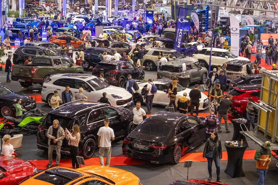 Mulți oameni vizitează un expoziție de mașini într-o hală mare cu diverse modele de autoturisme colorate și standuri de prezentare.