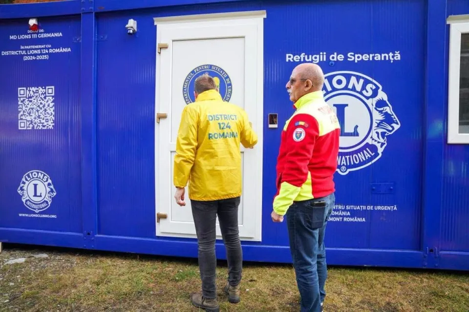 Doi bărbați stau în fața unei containere albastre pe care sunt scrise texte și un logo circular alb.