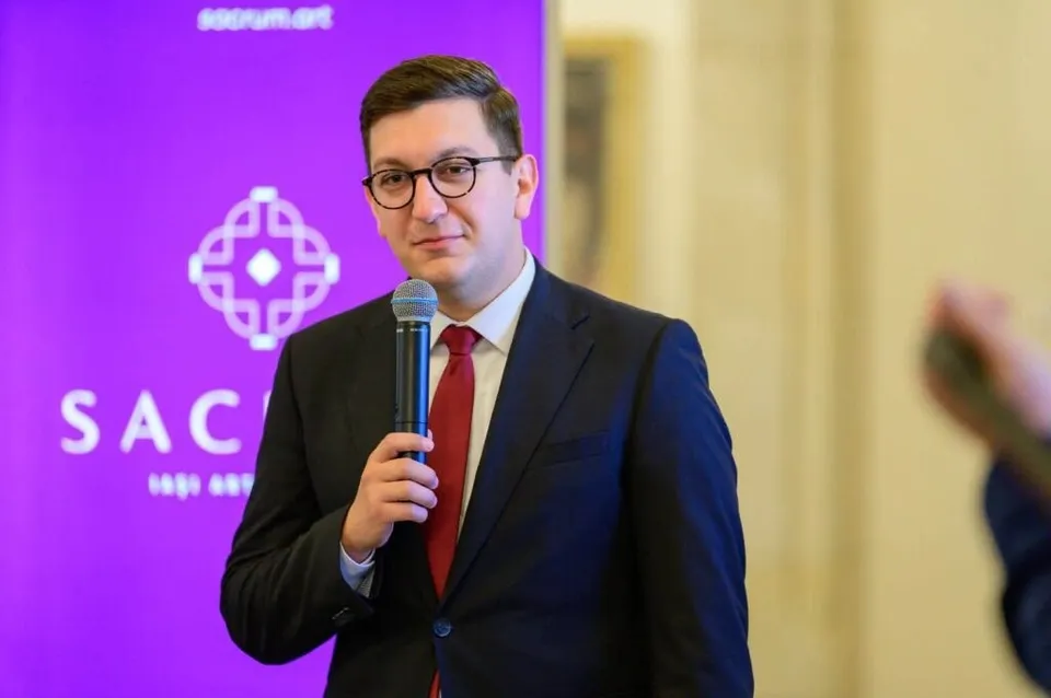Un bărbat cu ochelari și costum închis la culoare ține un microfon în mână în fața unui fundal mov cu un logo alb.