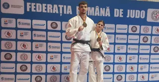Un bărbat și o femeie în costum de judo portând medalii aurii stau în fața unui panou albastru cu diverse logo-uri.