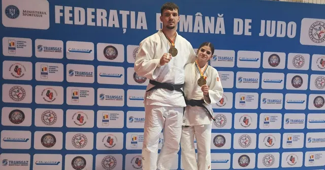 Un bărbat și o femeie în costum de judo portând medalii aurii stau în fața unui panou albastru cu diverse logo-uri.