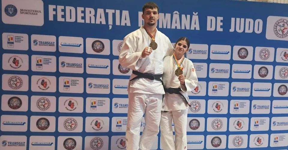 Un bărbat și o femeie în costum de judo portând medalii aurii stau în fața unui panou albastru cu diverse logo-uri.
