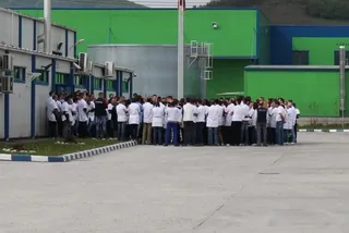 Un grup mare de persoane îmbrăcate în tricouri albe și pantaloni închizi stau adunate pe o suprafață asfaltată în fața unor clădiri industriale colorate.