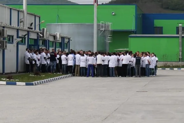 Un grup mare de persoane îmbrăcate în tricouri albe și pantaloni închizi stau adunate pe o suprafață asfaltată în fața unor clădiri industriale colorate.