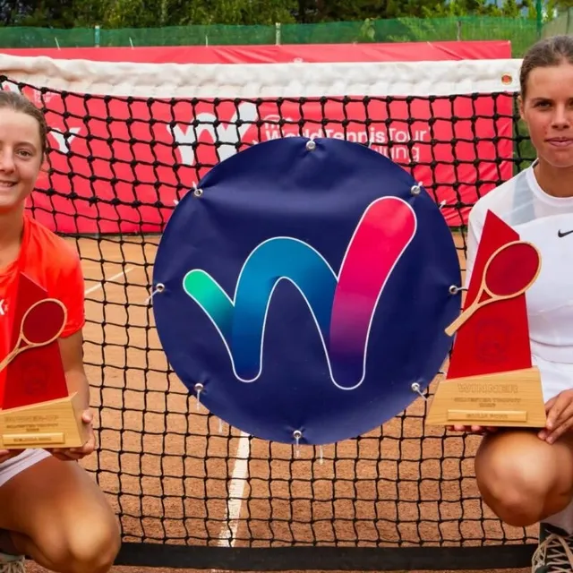 Două femei țin trofee cu o rachetă de tenis și un disc albastru cu un simbol colorat în fața unei țese de tenis.