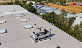 Un aparat de zbor fără pilot este poziționat pe un suport pe acoperișul unei clădiri industriale lângă un dispozitiv tehnic alb.