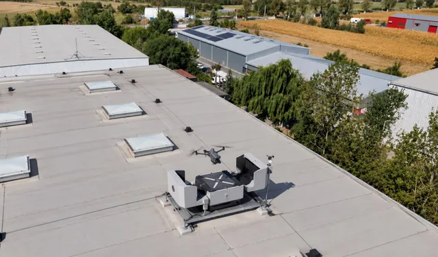 Un aparat de zbor fără pilot este poziționat pe un suport pe acoperișul unei clădiri industriale lângă un dispozitiv tehnic alb.