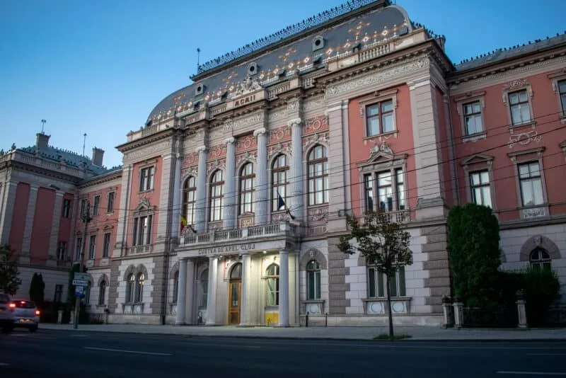 O clădire de stil arhitectural clasic cu coloane albe, ferestre înalte și fațadă roz este situată pe o stradă cu trotuar și un copac.
