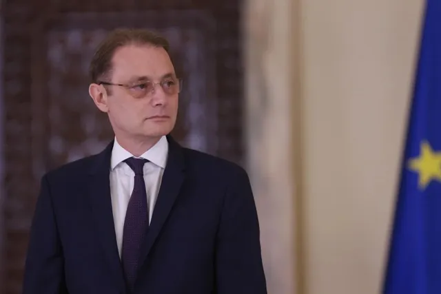 Un bărbat cu ochelari, purtând sacou închis la culoare, cămașă albă și cravată purpurie, stă în fața unui fundal neclar și a unei steaguri albastre.