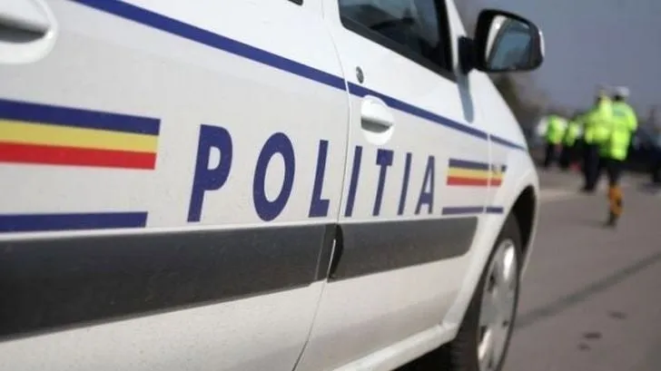 O mașină de poliție albă cu inscripția "POLIȚIA" și dungi tricolore pe lateral, cu un polițist în fundal.