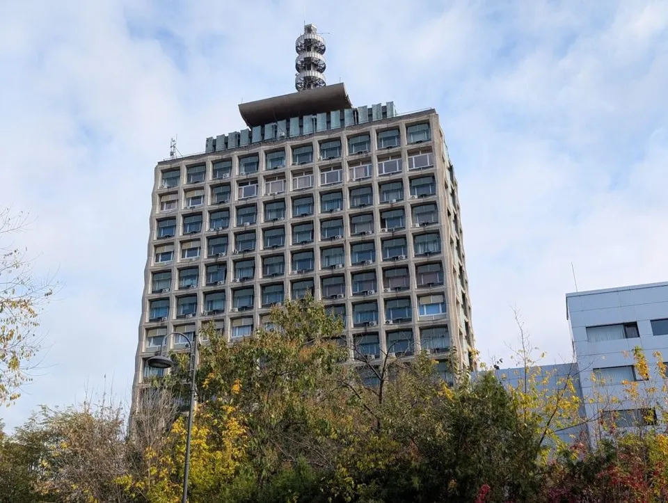 Un clădire înaltă de beton cu ferestre multiple este vizibilă sub un cer înnorat, parțial acoperită de copaci cu frunze verzi și galbene.