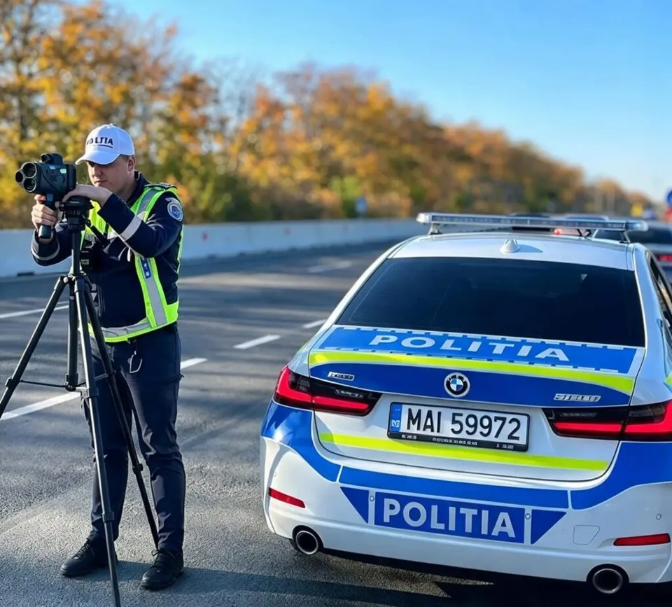Un polițist cu vestă reflectorizantă folosește un aparat de măsurat viteza montat pe un trepied lângă o mașină de poliție albastră și albă.