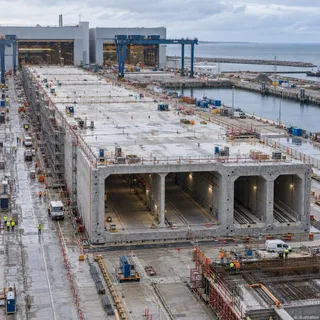 O structură de beton masivă cu mai multe secțiuni de tunel este în construcție lângă o zonă portuară cu macarale și utilaje de șantier.
