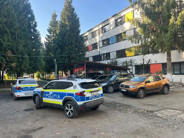Două mașini de poliție alb-albastre sunt parcate pe un teren de asfalt lângă o clădire cu ferestre colorate și copaci înalți.