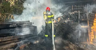 Un pompier cu cască roșie și echipament de protecție folosește un furtun pentru a stinge un incendiu lângă o structură de lemn carbonizat.
