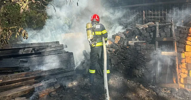 Un pompier cu cască roșie și echipament de protecție folosește un furtun pentru a stinge un incendiu lângă o structură de lemn carbonizat.