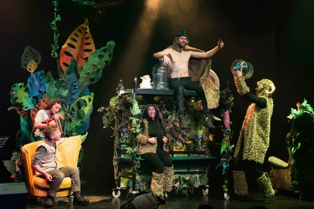 Patru actori costumați în personaje de teatru execută o scenă pe o platformă decorată cu plante și elemente de vegetație într-un decor întunecat.