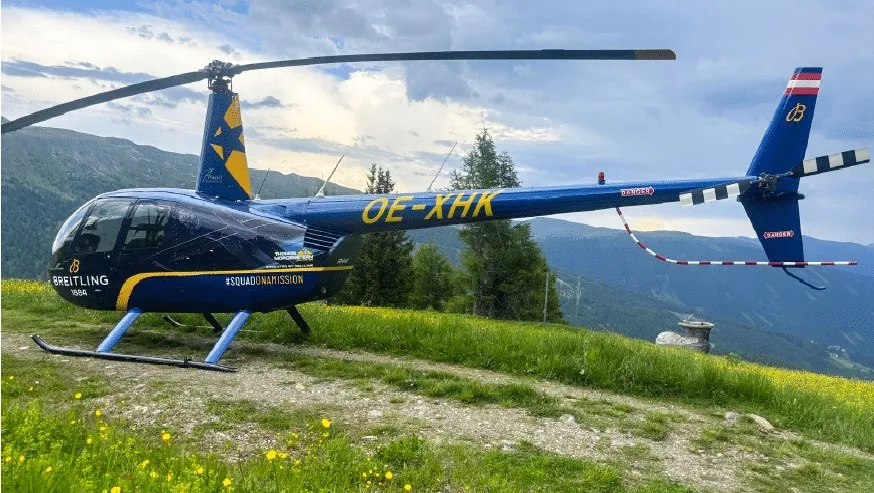 Un elicopter albastru cu marcaje galbene și textul OE-XHK este parcat pe o iarbă verde în fața unor munți sub un cer înnorat.