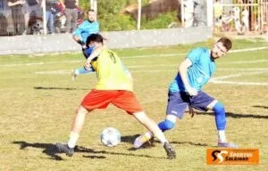 Un jucător în echipament galben și roșu se află pe teren de fotbal lângă un alt jucător în echipament albastru și o minge albă.