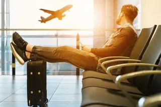 Un bărbat cu picioarele ridicate pe o valiză neagră stă întins pe un scaun de așteptare într-o incintă cu ferestre mari și un avion în fundal.