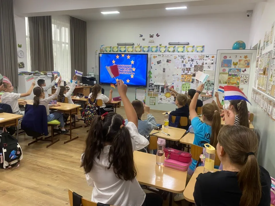 Elevi stau la bănci într-o sală de clasă, ridicând diverse obiecte și steaguri colorate în sus.