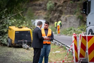 Un bărbat în costum negru stă lângă un bărbat cu vestă reflectorizantă portocalie lângă un echipament galben și bariere de circulație.