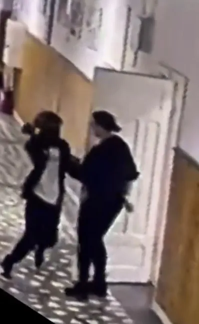 Două persoane îmbrăcate în haine închise stau în fața unei uși albe într-un coridor cu podea modelată.