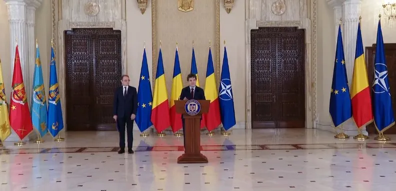 Două bărbați în costume întemeiate stau în fața unui podium de lemn, încadrați de mai multe steaguri colorate într-o încăpere cu podea de marmură.