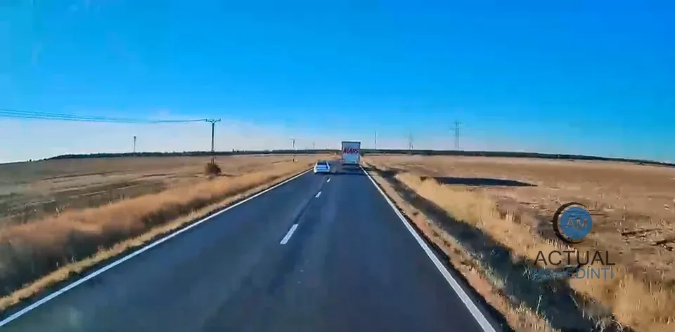 O stradă asfaltată cu o linie albă discontinuă separatoare trece printr-un câmp uscat sub un cer albastru, având un autoturism alb și un camion în depărtare.