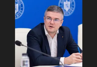 Un bărbat cu ochelari și sacou închis la culoare stă la o masă cu un microfon în față și un pix în mână.