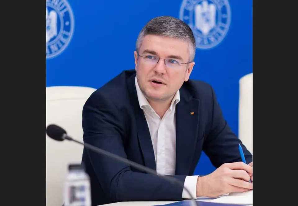 Un bărbat cu ochelari și sacou închis la culoare stă la o masă cu un microfon în față și un pix în mână.